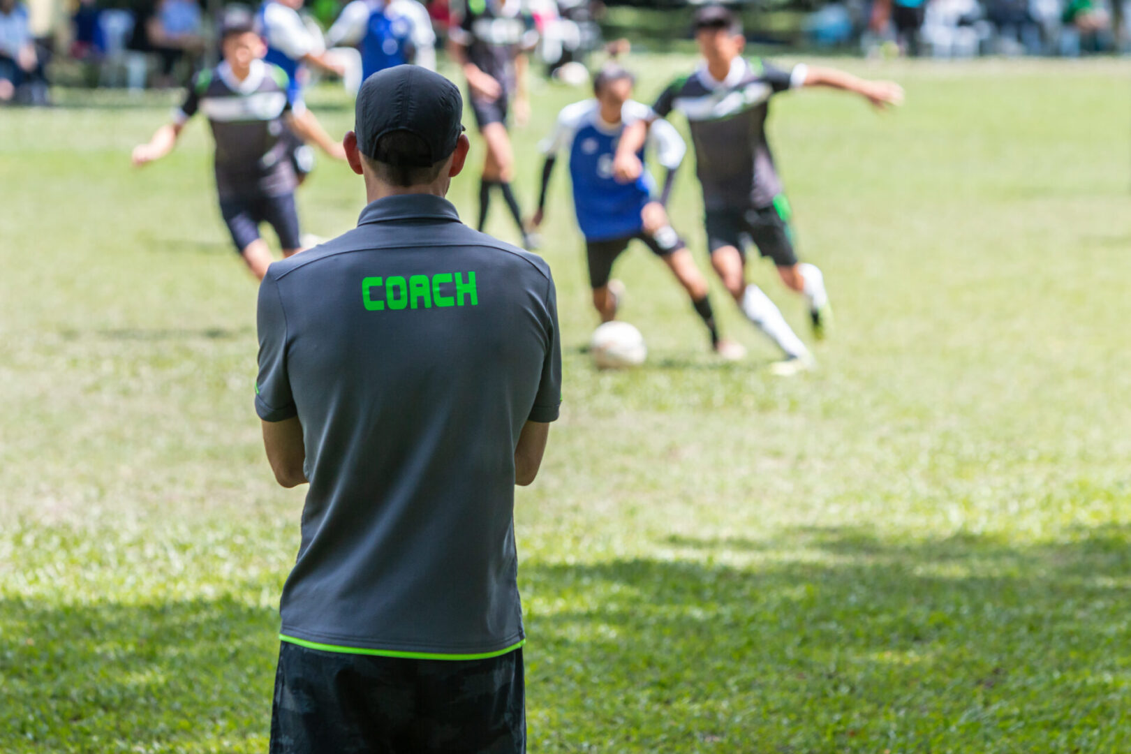 Male soccer or football coach in gray shirt with word COACH written on back, standing on the sideline watching his team play, good for sport or coaching concept