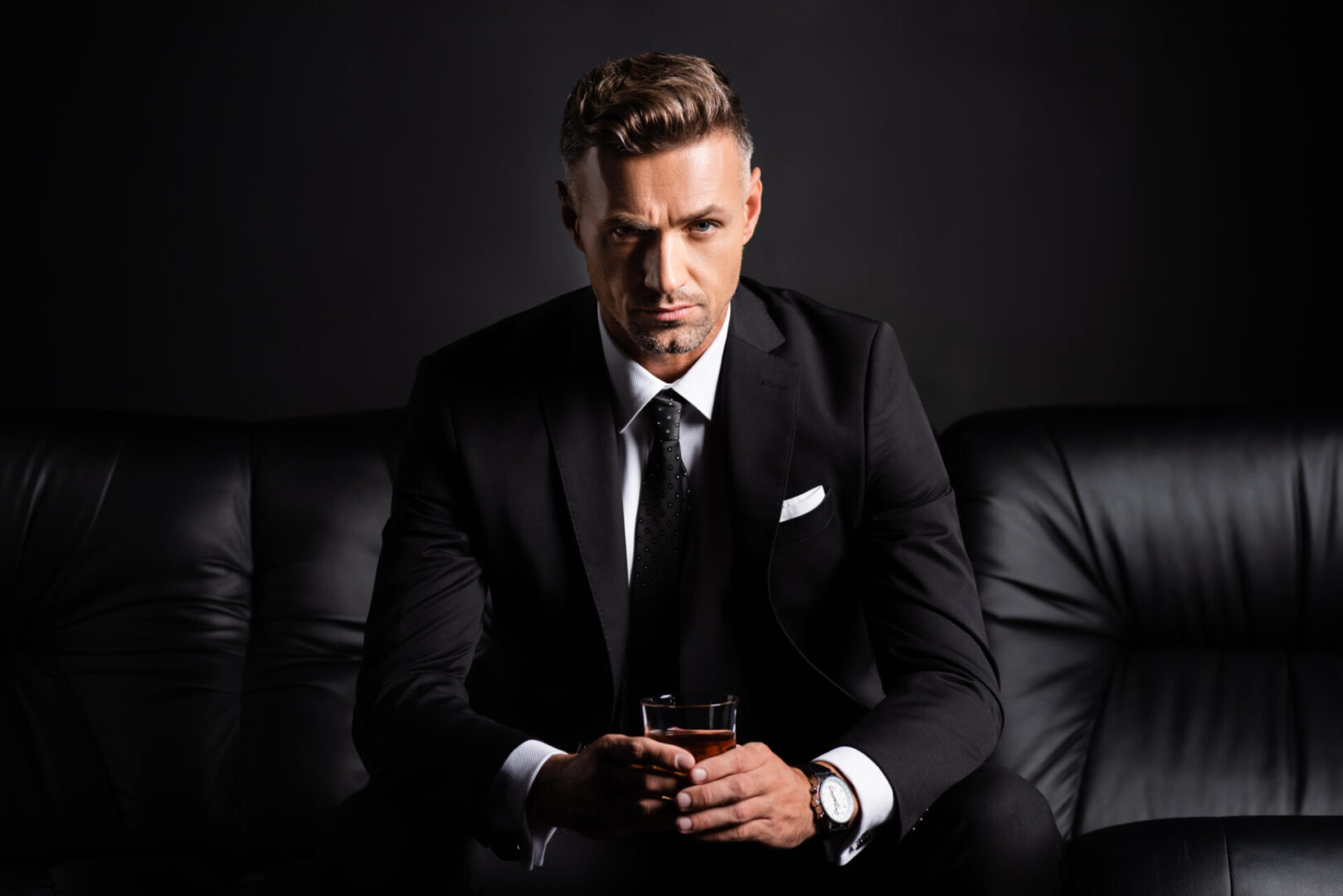 Handsome businessman with glass of whiskey looking at camera on couch isolated on black